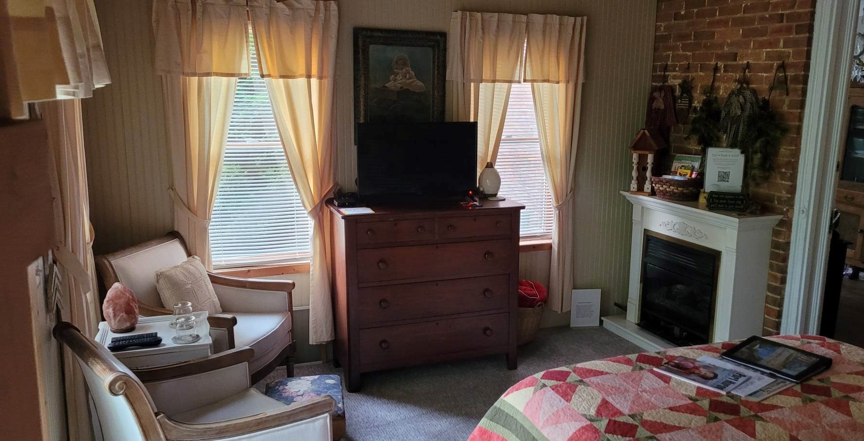 A cozy bedroom sitting area featuring a white fireplace mantel against an exposed brick wall, a dark wood dresser holding a television between two windows, two armchairs in the corner, and the edge of a bed with a patchwork quilt.