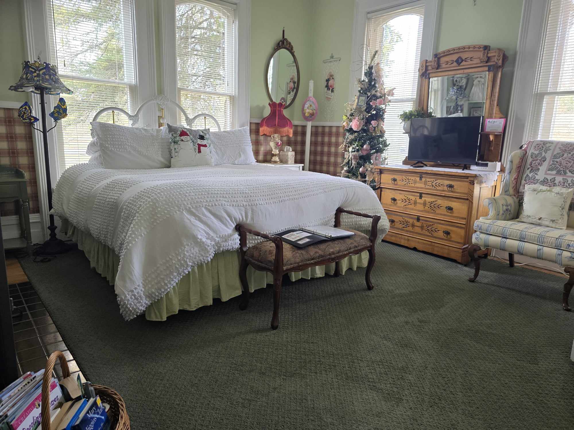 A spacious bedroom featuring a large bed with white linens, a wooden dresser with a mirror, a wingback chair, and a Christmas tree decorated in the corner.