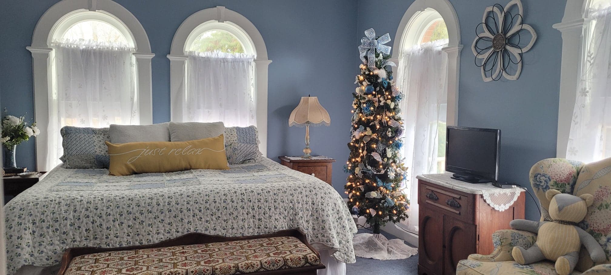 A bedroom with light blue walls and three distinct arched windows, featuring a bed with a white patterned quilt and yellow lumbar pillow, a slim decorated Christmas tree, and a teddy bear sitting on a wingback chair.