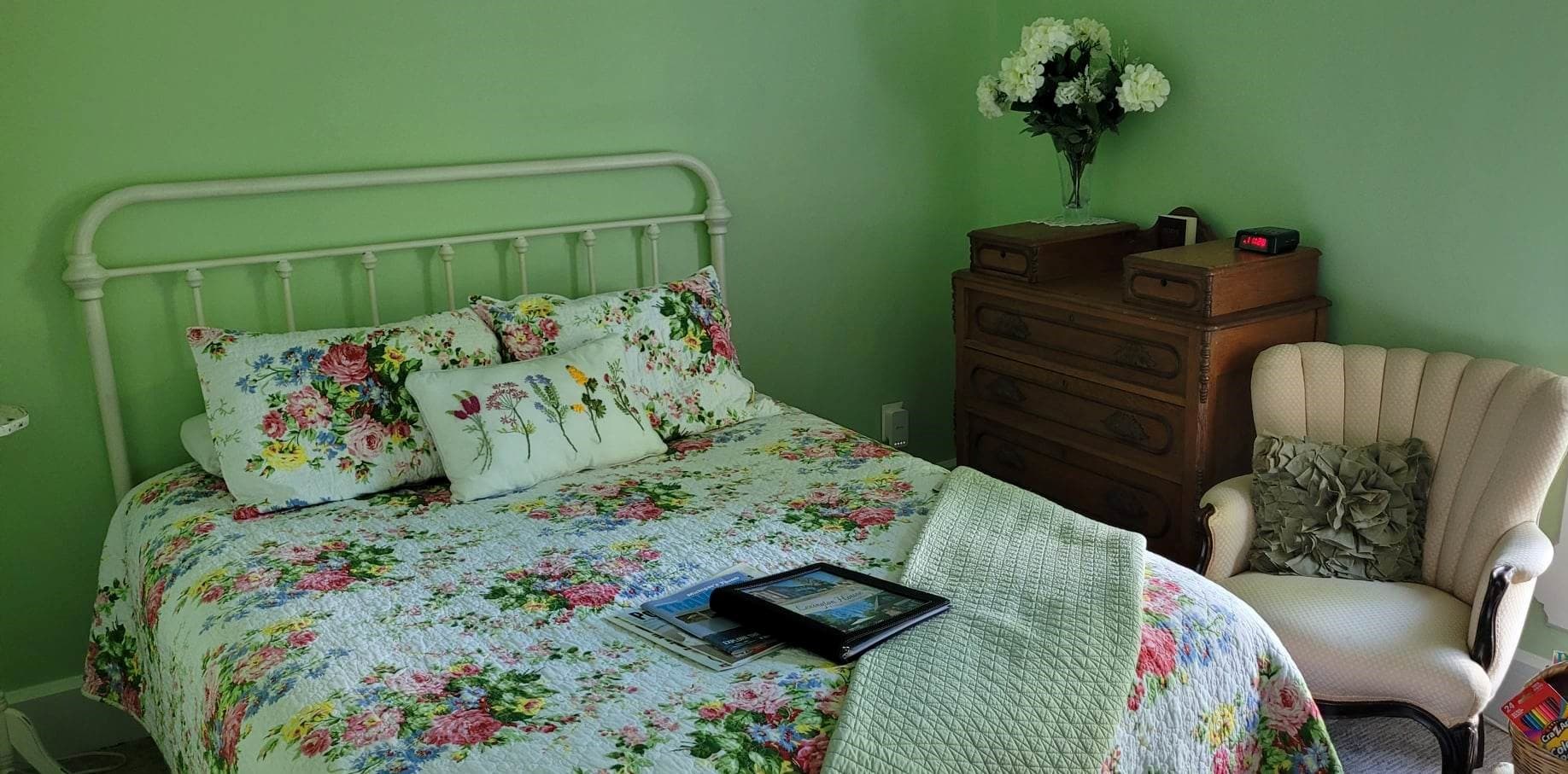 A bright bedroom with soft green walls featuring a green iron farmhouse-style bed dressed in floral bedding. Next to the bed is a wooden chest of drawers topped with a vase of white flowers, and a cream-colored armchair with an Eiffel Tower pillow sits in the corner.