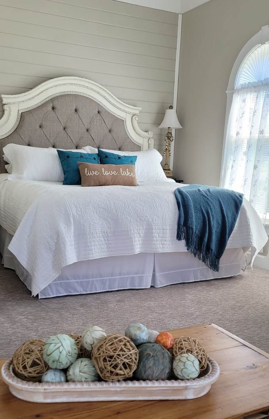 A bright bedroom featuring a large, elegant headboard with beige tufting and a white frame. The bed is dressed in white linens with teal accent pillows and a matching teal throw blanket. In the foreground, a wooden tray filled with decorative yarn balls sits on a table.