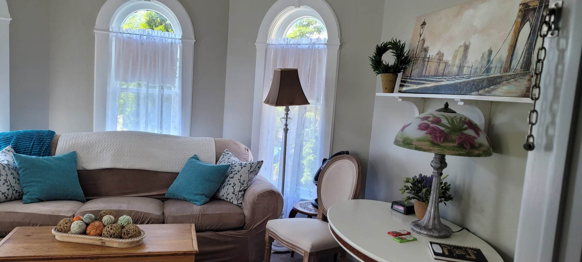 A cozy sitting room featuring two arched windows with sheer curtains behind a beige sofa accented with teal pillows. A wooden coffee table with decorative spheres sits in the center, and a small desk area with a lamp and painting is visible on the right.