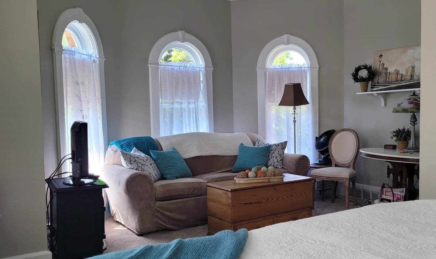 A cozy sitting area featuring three distinct arched windows with sheer curtains behind a beige sofa accented with teal throw pillows. A wooden chest serves as a coffee table, and a vintage sewing machine table sits to the right.