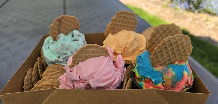 A box of colorful ice cream scoops with waffle cones on top, set on a rustic table by a green landscape.