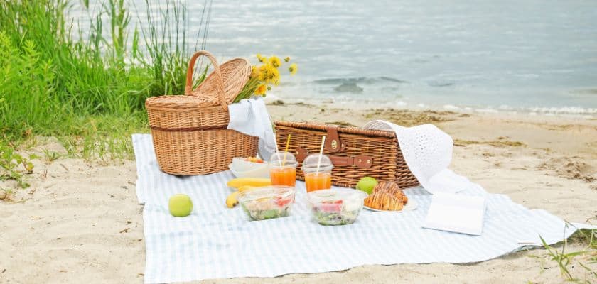 A picnic setup on the beach featuring a wicker basket, food containers, and drinks on a checkered blanket.