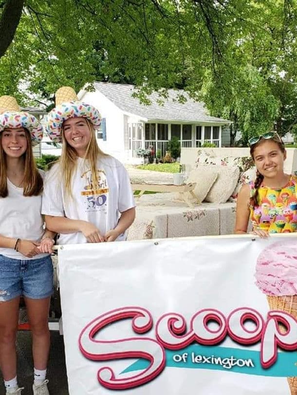 Three young women wearing colorful hats pose with a banner for "Scoop of Lexington" at an outdoor event.