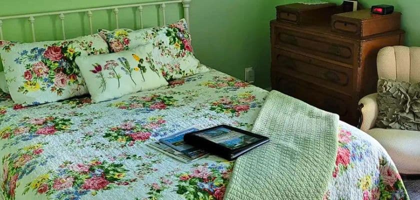 A floral-themed bed with a decorative pillow, a throw blanket, and magazines on top, accompanied by vintage furniture.