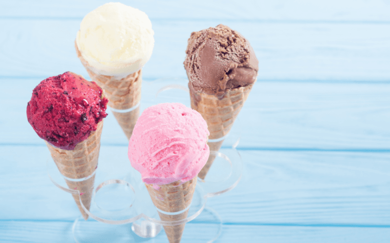 Four ice cream cones in a holder, featuring flavors of raspberry, strawberry, vanilla, and chocolate on a blue background.