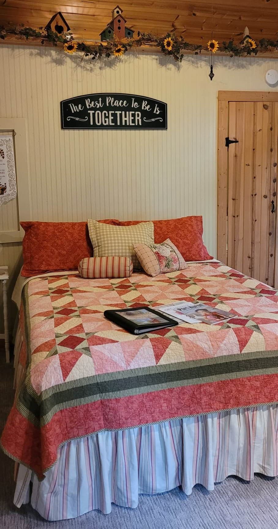 A cozy, rustic bedroom featuring a bed with a pink and red star-patterned quilt and a black sign hanging above the headboard that reads 'The Best Place to be is TOGETHER'.